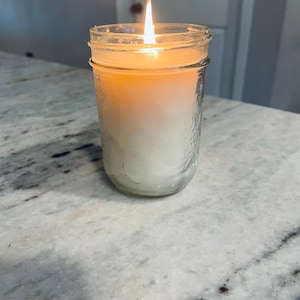 A lit white candle in a clear mason jar with a textured surface sits on a speckled white countertop, with a blurred light-colored door in the background.