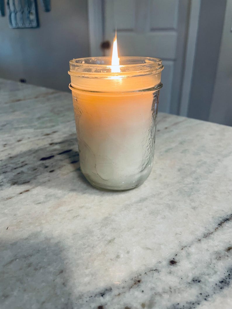 A lit white candle in a clear mason jar with a textured surface sits on a speckled white countertop, with a blurred light-colored door in the background.