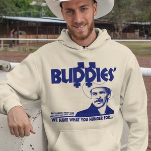 May include: A beige hoodie with a blue graphic print of a cowboy wearing a hat and the text "Buddies' Restaurant/Party Bar 5742 North Clark Street, Chicago WE HAVE WHAT YOU HUNGER FOR..."