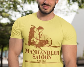 Può includere: Una maglietta gialla con una grafica marrone di un cowboy a cavallo. La grafica include il testo "Manhandler Saloon Chicago".