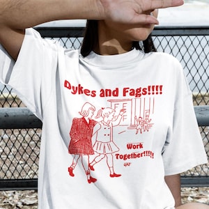 May include: White t-shirt with red text and graphic. The text reads "Dykes and Fags!!!!" above an illustration of two figures walking together. Below the illustration is the text "Work Together!!!!"