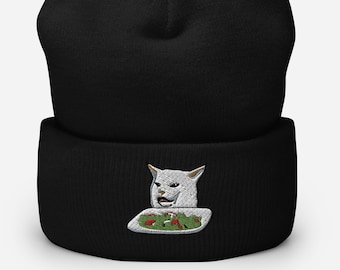 Pode incluir: Boné preto com um gato branco bordado na frente. O gato está olhando para um prato de salada e tem uma expressão de surpresa no rosto.