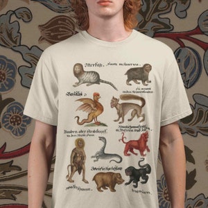 Könnte beinhalten: Cremefarbenes T-Shirt mit einer Vintage-Illustration von Fabelwesen und Text in klassischer Schrift. Das Design umfasst eine Vielzahl von fantastischen Wesen, wie eine katzenartige Kreatur mit Löwengesicht, einen geflügelten Hahn und ein bärenähnliches Wesen.