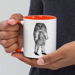 May include: A white ceramic mug with an orange rim features a black and white illustration of a cat standing upright with its arms crossed. The cat is wearing pants and shoes.