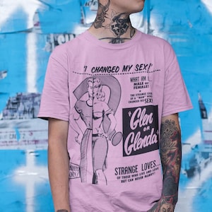 May include: A light pink short-sleeved t-shirt featuring a vintage-style graphic print. The design includes the text "I CHANGED MY SEX!", "Glen or Glenda", and "STRANGE LOVES...", alongside a split illustration of a person in a dress.