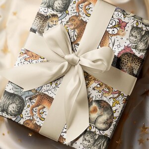May include: A square gift box wrapped in patterned paper featuring illustrations of various cat-like creatures in shades of brown, gray, and cream. A cream-colored satin ribbon and bow adorn the top, creating a luxurious presentation.