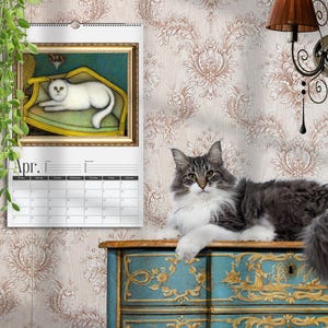 May include: A wall calendar for April featuring a framed painting of a white cat resting on a yellow chair. A long-haired gray and white cat sits on a blue and gold painted wooden chest. The calendar has a white background with black text.