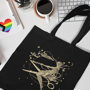 May include: A black tote bag featuring a gold design of a woman seated on a crescent moon, surrounded by stars and clouds. The woman is wearing a flowing dress and has long hair.