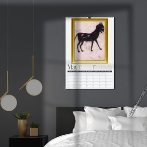 May include: A wall calendar for the month of May featuring a framed illustration of a black cat with small red and blue spots on its side. The calendar has a white background with a black grid for dates and the text May printed at the top.