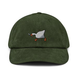 Puede incluir: Una gorra de béisbol de pana verde con una imagen bordada de un ganso blanco sosteniendo un cuchillo.
