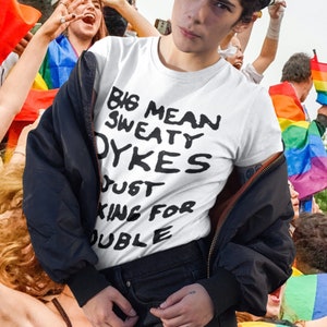 May include: A white t-shirt with black text that reads "Big Mean Sweaty Dykes Just King For Double". The person wearing the shirt is standing in front of a crowd of people holding rainbow flags.