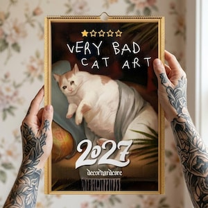 May include: A 2027 wall calendar with a gold frame featuring an illustration of a sad white and orange cat lying on a blue pillow. The text reads VERY BAD CAT ART with one gold star and the word decorhardcore at the bottom in white lettering.