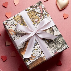 May include: A wrapped gift box with a light pink ribbon and bow. The wrapping paper features a whimsical design of cats and tigers in various poses. Scattered around the box are paper hearts in shades of red and pink, and a gold heart.
