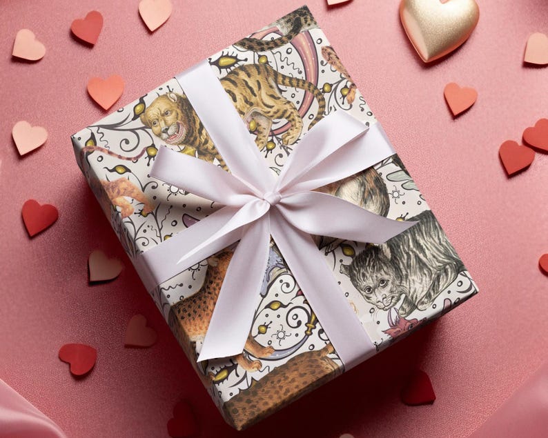 May include: A wrapped gift box with a light pink ribbon and bow. The wrapping paper features a whimsical design of cats and tigers in various poses. Scattered around the box are paper hearts in shades of red and pink, and a gold heart.