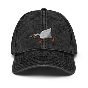 Puede incluir: Una gorra de béisbol de algodón vintage lavada en negro con una imagen bordada de un ganso blanco que sostiene un cuchillo en su pico.
