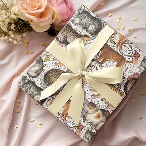 May include: A wrapped gift box with a cream-colored ribbon. The wrapping paper features a pattern of various cat illustrations in shades of brown, gray, and white. The background is a soft pink fabric, with a bouquet of flowers in the upper left corner.