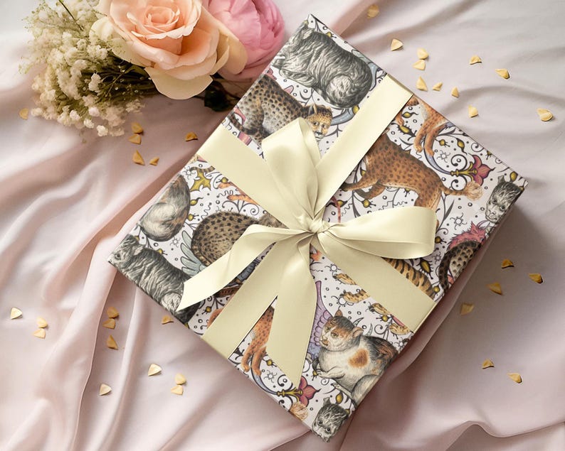 May include: A wrapped gift box with a cream-colored ribbon. The wrapping paper features a pattern of various cat illustrations in shades of brown, gray, and white. The background is a soft pink fabric, with a bouquet of flowers in the upper left corner.
