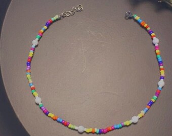 May include: A colourful beaded necklace with white beads interspersed throughout. The necklace is made of small, round beads in a variety of colours, including red, orange, yellow, green, blue, and purple.