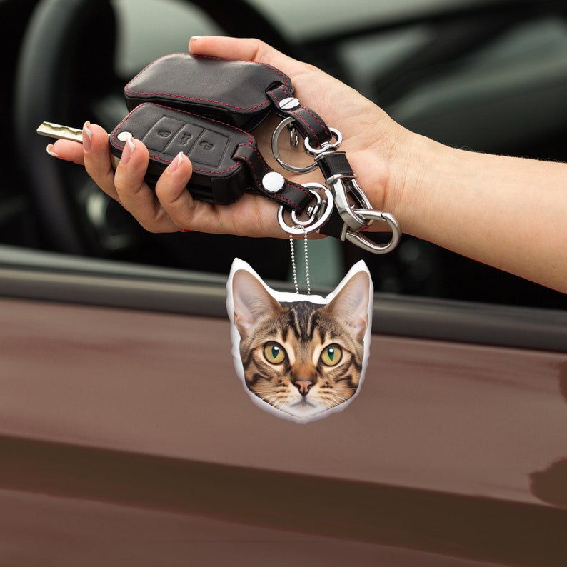 K&ouml;nnte beinhalten: Ein Autoschl&uuml;sselanh&auml;nger und Schl&uuml;ssel mit einem schwarzen Lederetui und roten N&auml;hten. Ein katzenf&ouml;rmiger Lufterfrischer mit einem braunen und beigen Tabby-Muster h&auml;ngt an den Schl&uuml;sseln. Die Katze hat gelbe Augen und einen wei&szlig;en Rand.