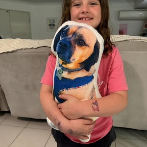 K&ouml;nnte beinhalten: Ein individuell bedrucktes, hundef&ouml;rmiges Kissen. Das Kissen zeigt ein Foto eines Hundes mit braunem und schwarzem Fell, der ein blaues Geschirr und einen Anh&auml;nger tr&auml;gt. Das Kissen wird von einer Person gehalten, die ein rosa T-Shirt tr&auml;gt.
