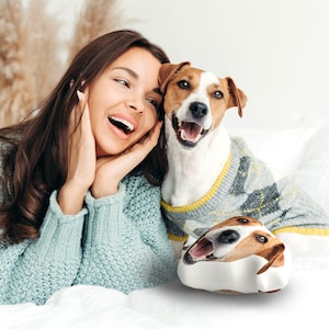 K&ouml;nnte beinhalten: Eine Frau und ein Jack Russell Terrier-Hund l&auml;cheln. Der Hund tr&auml;gt einen grauen Pullover mit gelbem Besatz. Ein wei&szlig;es Kissen mit dem Gesicht des Hundes ist im Vordergrund. Die Frau tr&auml;gt einen hellblauen Pullover.