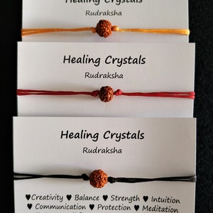 May include: Three bracelets with brown beads and colourful strings. The bracelets are labelled "Healing Crystals Rudraksha". The bottom bracelet has a list of benefits associated with the beads, including creativity, balance, strength, intuition, communication, protection, meditation, stress relief, focus, wisdom, joy, self healing, luck and good fortune, grief and mourning, and anxiety relief.