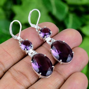 May include: A pair of silver-toned drop earrings featuring oval and round, deep purple gemstones. The earrings have a lever-back closure and are set in a decorative silver metal setting. The gemstones are a rich, dark purple color.