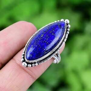 May include: A silver ring featuring a large oval-shaped lapis lazuli gemstone. The gemstone is a deep blue colour with gold flecks throughout.