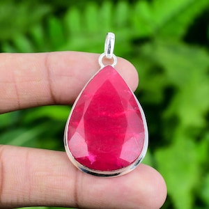 May include: A teardrop-shaped pendant featuring a faceted, vibrant pinkish-red gemstone set in a polished silver-toned metal frame. The pendant includes a small silver-toned bail at the top for attachment, showcasing a clear, geometric cut.