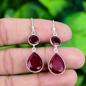 Könnte beinhalten: Silberne Ohrhänger mit zwei tiefroten Edelsteinen. Der obere Stein ist rund, der untere tropfenförmig. Die Ohrringe hängen an einem silbernen Haken. Die Edelsteine sind ein sattes, dunkles Rot.