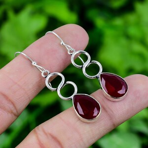 May include: Silver dangle earrings with a teardrop-shaped red gemstone. The earrings feature a twisted, infinity-like design.