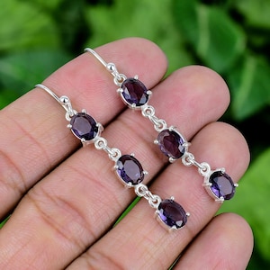 May include: A pair of silver drop earrings featuring oval, deep purple gemstones. Each earring has three gemstones connected by small silver links. The earrings are displayed against a blurred green background.