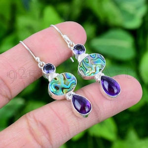 May include: Sterling silver earrings featuring abalone shell and amethyst gemstones. The earrings have a teardrop-shaped amethyst gemstone hanging from a round abalone shell. The abalone shell is surrounded by a smaller amethyst gemstone.