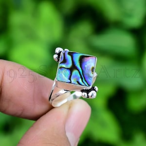 May include: A silver ring featuring a square abalone shell inlay. The shell displays iridescent colours of blue, green, and purple.