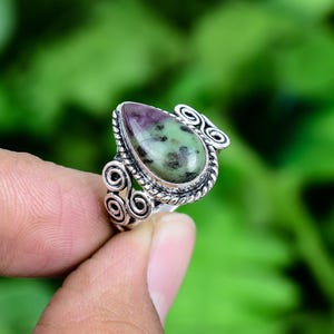 May include: A silver ring featuring a teardrop-shaped zoisite stone with green and purple hues. The metal band displays a twisted rope border around the stone and decorative scrollwork patterns on the sides, creating a detailed metal setting.