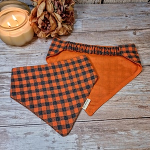Könnte beinhalten: Zwei Hunde-Bandanas, eines dreieckig und eines rechteckig, aus orange-schwarz kariertem Stoff. Das rechteckige Bandana hat einen elastischen Hals. Die Bandanas liegen auf einer Holzoberfläche, mit einer Kerze und Blumen im Hintergrund.