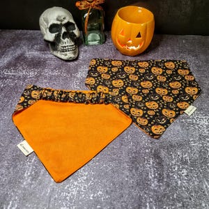 May include: Two orange and black dog bandanas. One bandana has a black background with orange pumpkins and the other bandana is solid orange. Both bandanas have a ruffled edge.