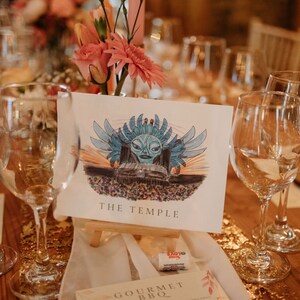 May include: A table setting with a white card featuring a blue and green illustration of a temple with the text "THE TEMPLE". The card is on a wooden easel and surrounded by wine glasses and pink flowers.