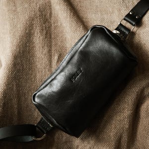 Puede incluir: Riñonera de cuero negro con cierre de cremallera y correa ajustable. La bolsa tiene la palabra "POLEY" grabada en relieve en la parte delantera. La bolsa es rectangular y tiene una cremallera plateada.