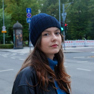 Op de afbeelding: Een vrouw met lang bruin haar die een donkerblauwe muts, een donkerblauwe jas en een lichtblauw shirt draagt.