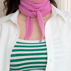 May include: A pink crocheted scarf tied around the neck of a person wearing a white button-down shirt and a green and white striped shirt.