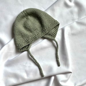 May include: A sage green knitted baby hat with ties. The hat is made of soft yarn and has a simple design.