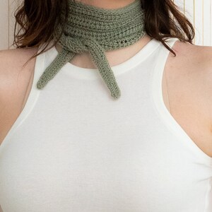 May include: A sage green crocheted neck scarf tied in a knot around the neck. The scarf is made of a soft, textured yarn.