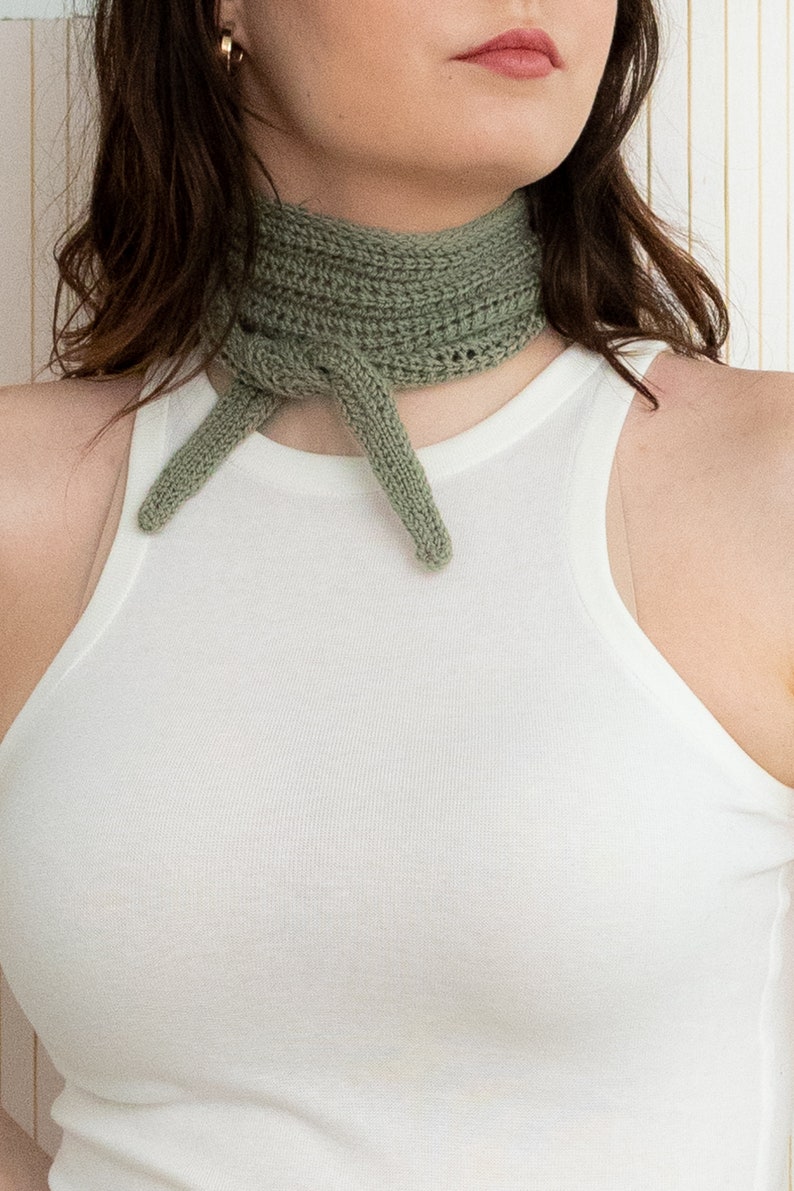 May include: A sage green crocheted neck scarf tied in a knot around the neck. The scarf is made of a soft, textured yarn.