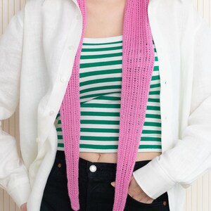 May include: A pink crocheted scarf worn around the neck of a person wearing a white button-down shirt and black jeans.