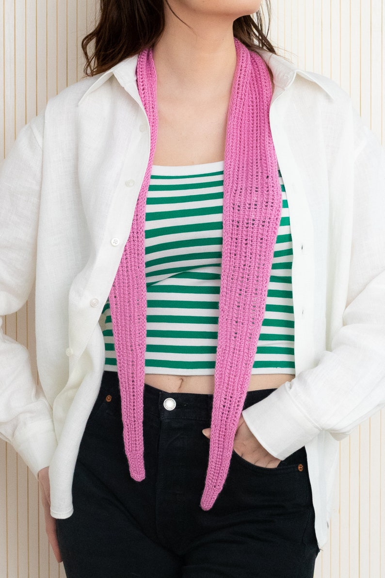 May include: A pink crocheted scarf worn around the neck of a person wearing a white button-down shirt and black jeans.