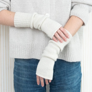 May include: A pair of white knitted fingerless gloves with a ribbed texture. The gloves are worn on the hands of a person wearing a light gray sweater and blue jeans.