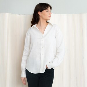 Peut inclure: Une chemise blanche en lin avec des manches longues et un col classique. La chemise est partiellement déboutonnée. Le modèle porte un pantalon noir. L'arrière-plan est un motif à rayures verticales beige clair.