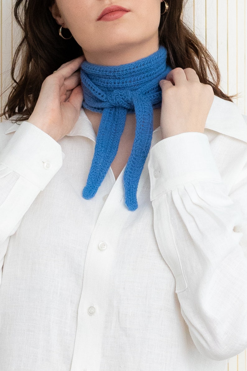 May include: A woman wearing a white button-down shirt with a blue knitted scarf tied around her neck.