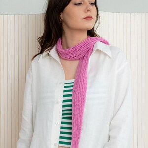 May include: A woman wearing a white linen shirt with a pink crocheted scarf. The scarf is long and hangs down to her waist.